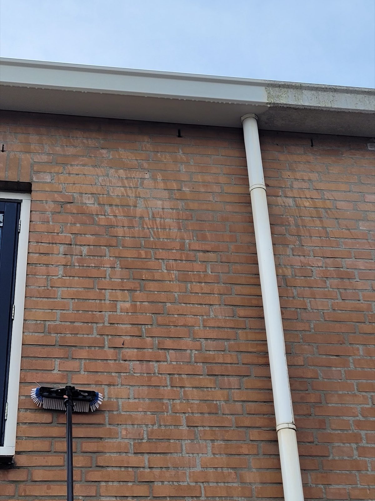 Witte dakgoot en regenpijp langs een bakstenen muur, met een reinigingsborstel
